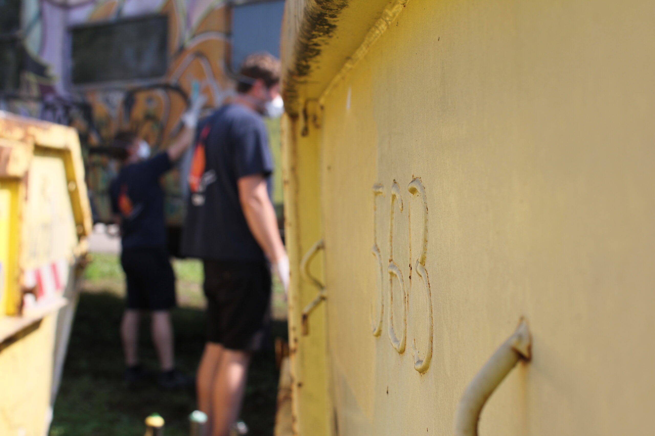 Gelbe Wand mit eingeritzten Zeichen, im Hintergrund Personen in schwarzer Kleidung.