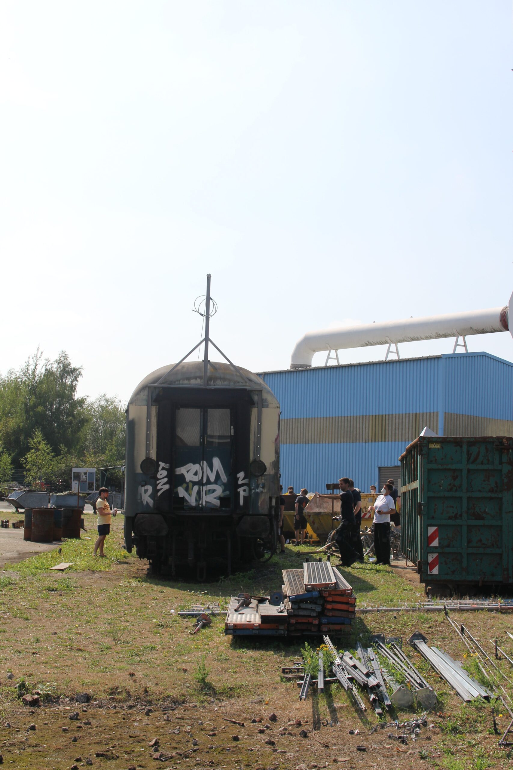 Alte Eisenbahnwaggons und Industrieanlagen auf einem Gelände mit mehreren Personen.