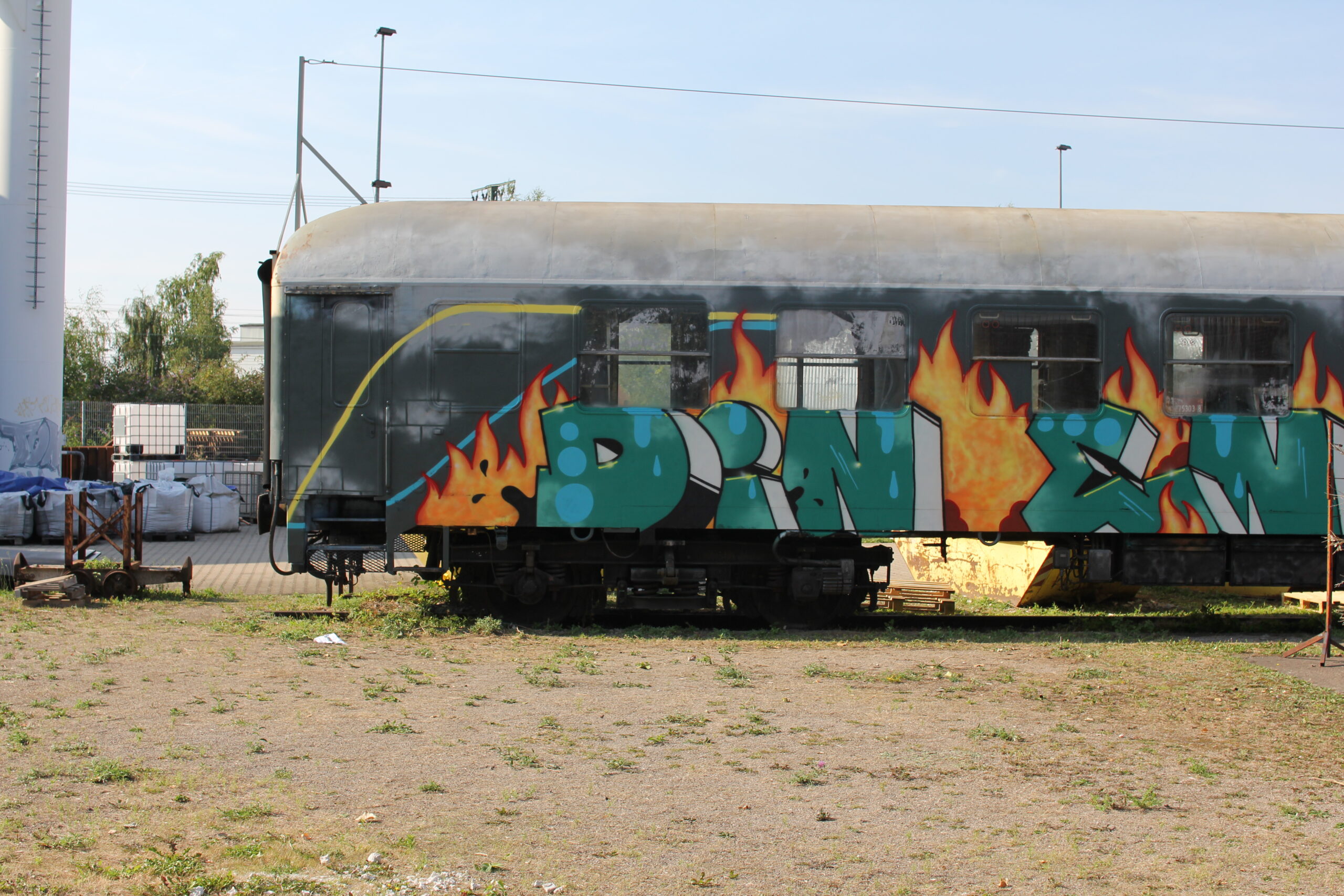 Stillgelegter Zugwagen mit großflächigem Graffiti in Grün und roten Flammen auf Industriegelände.