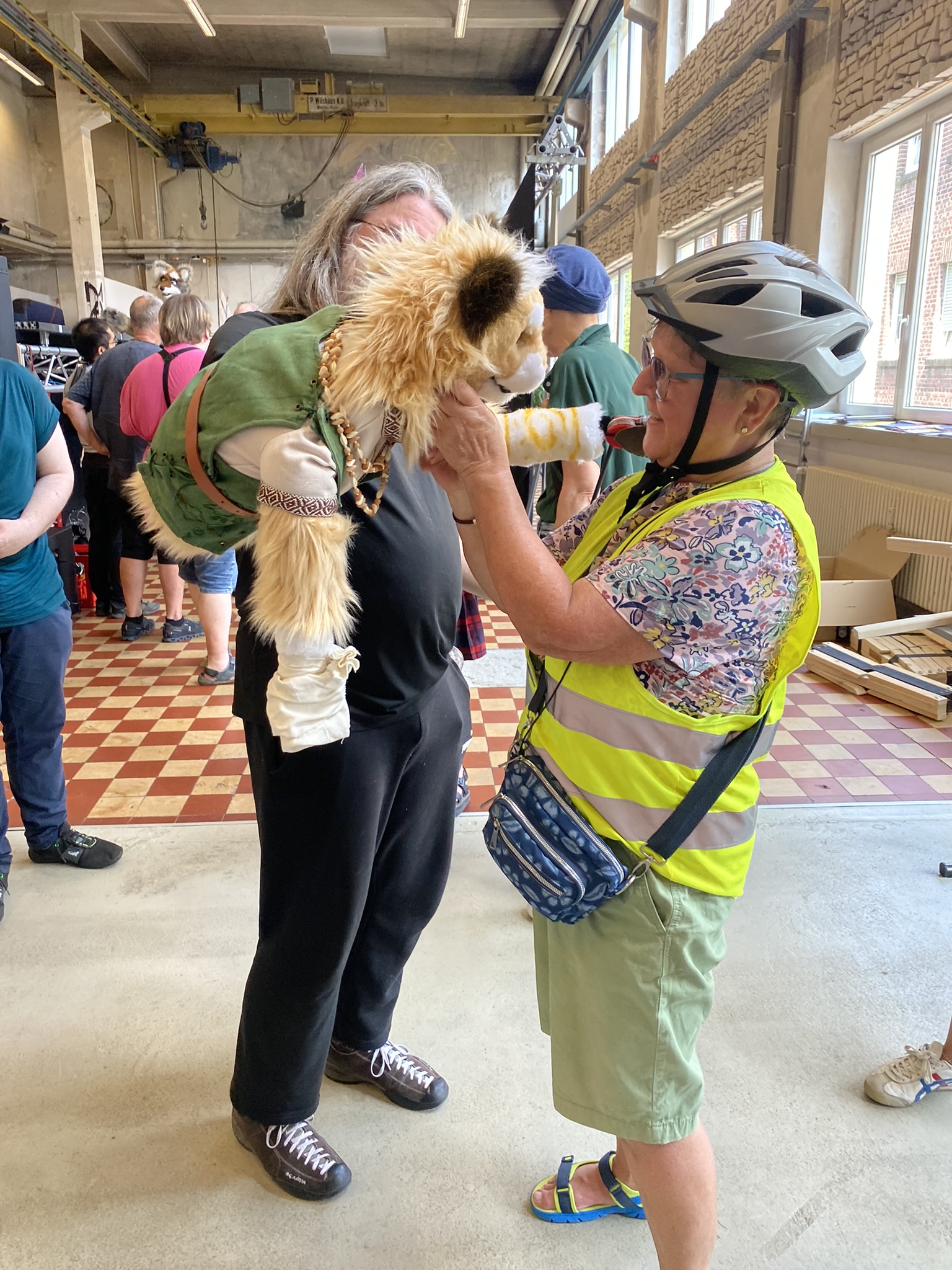 Eine Frau mit grauem Haar hält einen Hund in grüner Jacke. Ein Mann im Helm und Warnweste steht daneben.