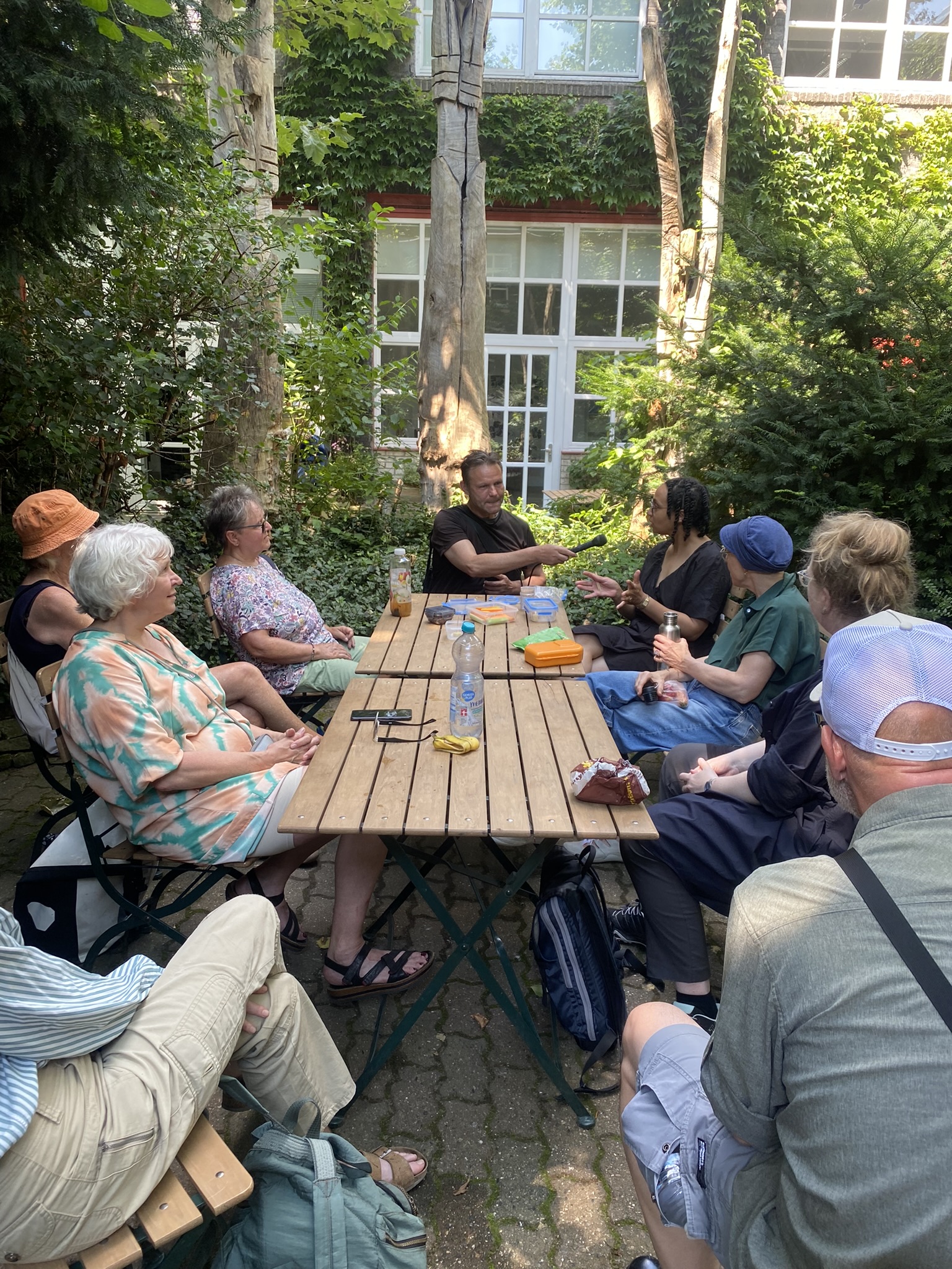 Etwa zehn Personen sitzen an Holztischen in einem begrünten Innenhof und diskutieren miteinander.
