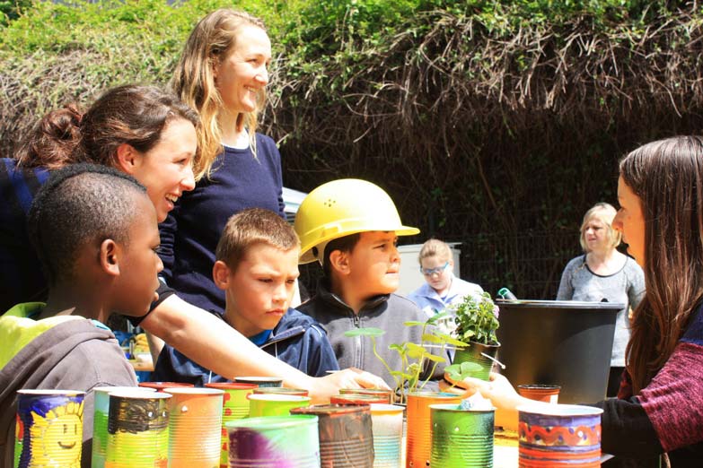 Gruppe von Kindern und Erwachsenen bei einem Gartenprojekt mit Pflanzen und recycelten Behältern.
