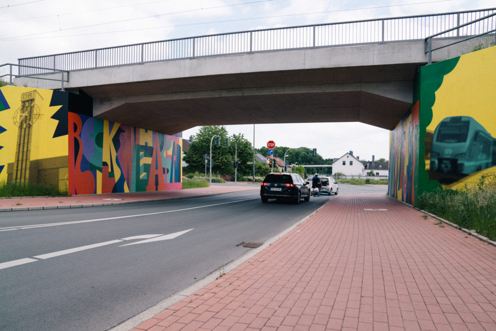Unterführung mit buntem Graffiti, Straße, Gehweg und parkenden Autos darunter.