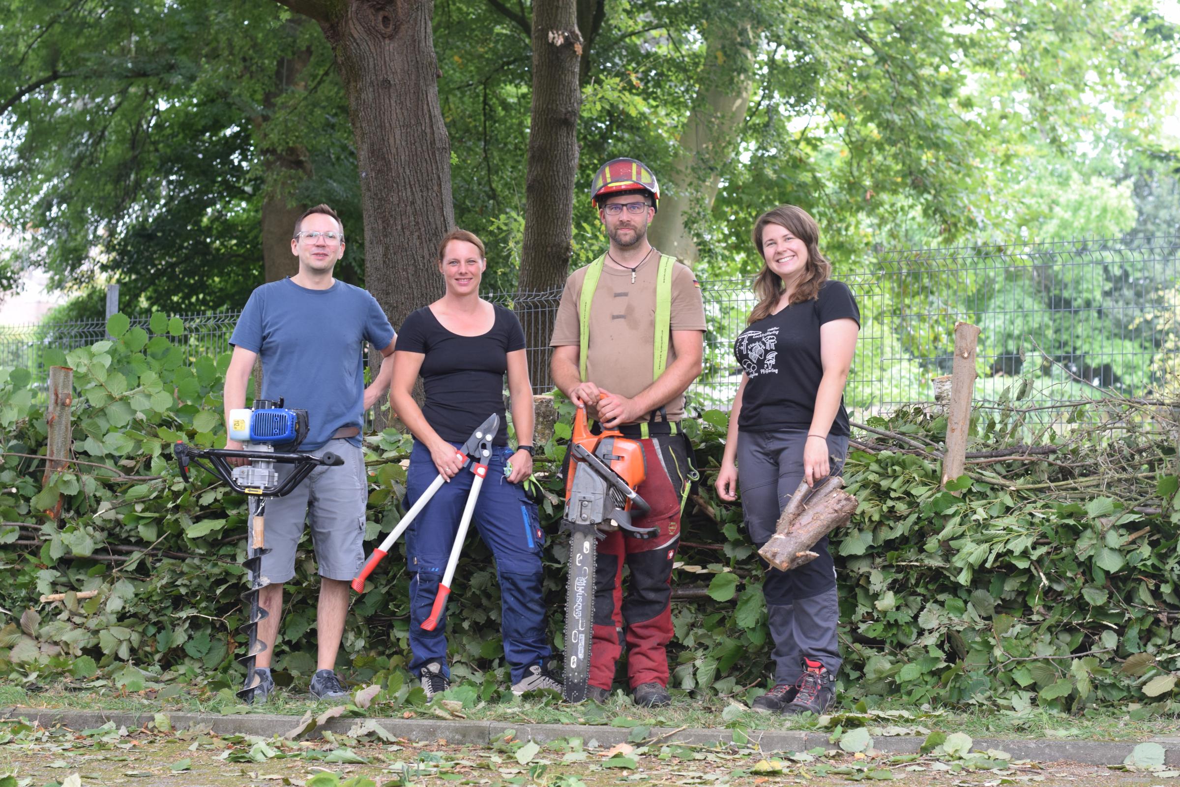 Vier Personen mit Gartengeräten vor einer Benjeshecke