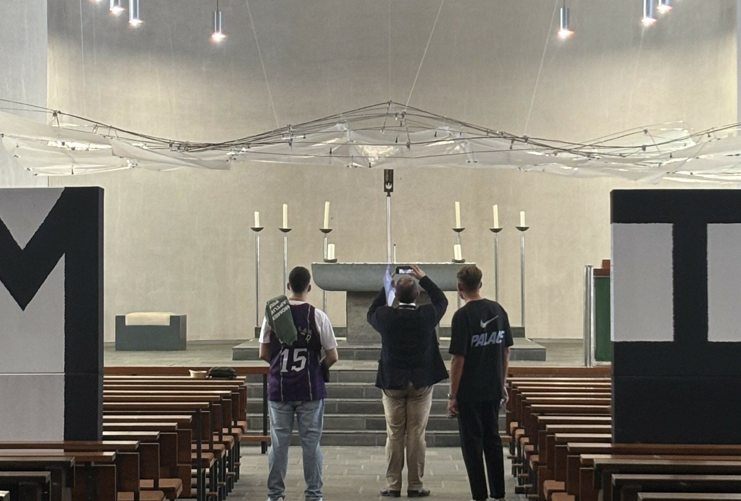 Drei Personen stehen in einer modernen Kirche vor einem Altar mit Kerzenständern.