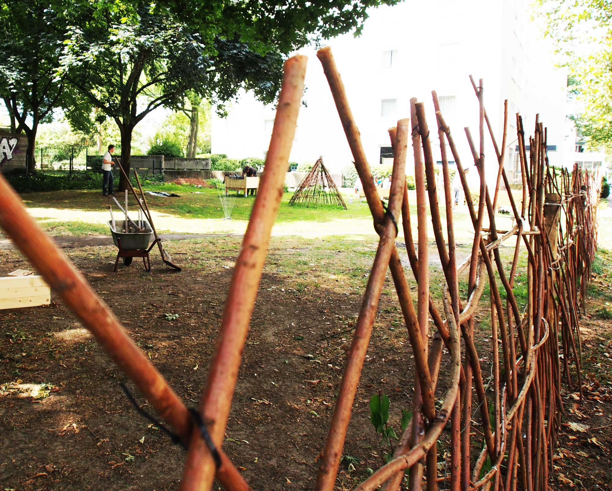 Gemeinschaftsgarten mit Weidenzaun, Hochbeeten und Spielgeräten unter Bäumen.