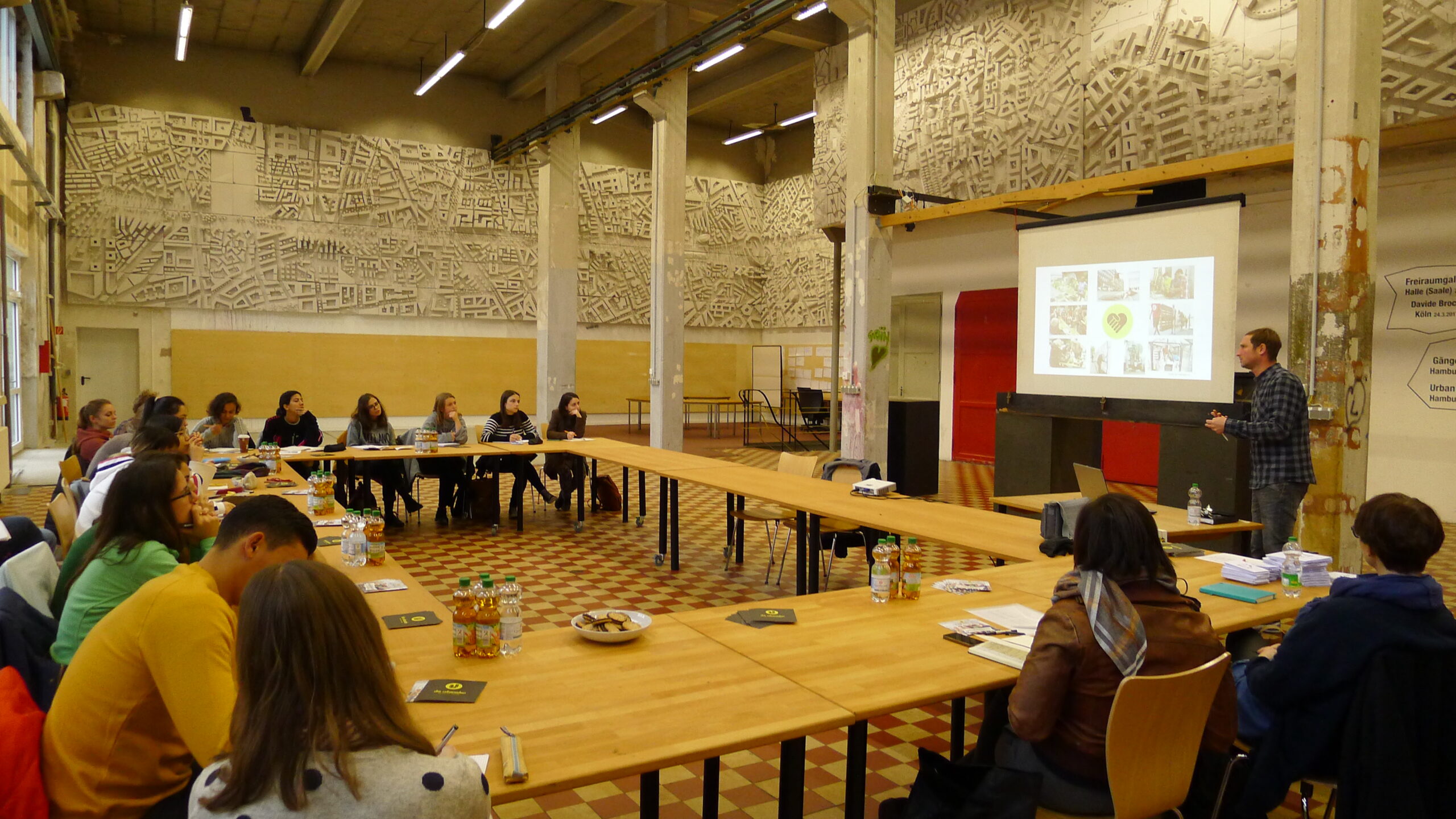 Eine Veranstaltung in einer Halle mit Teilnehmern an Tischen und Präsentation an Leinwand.