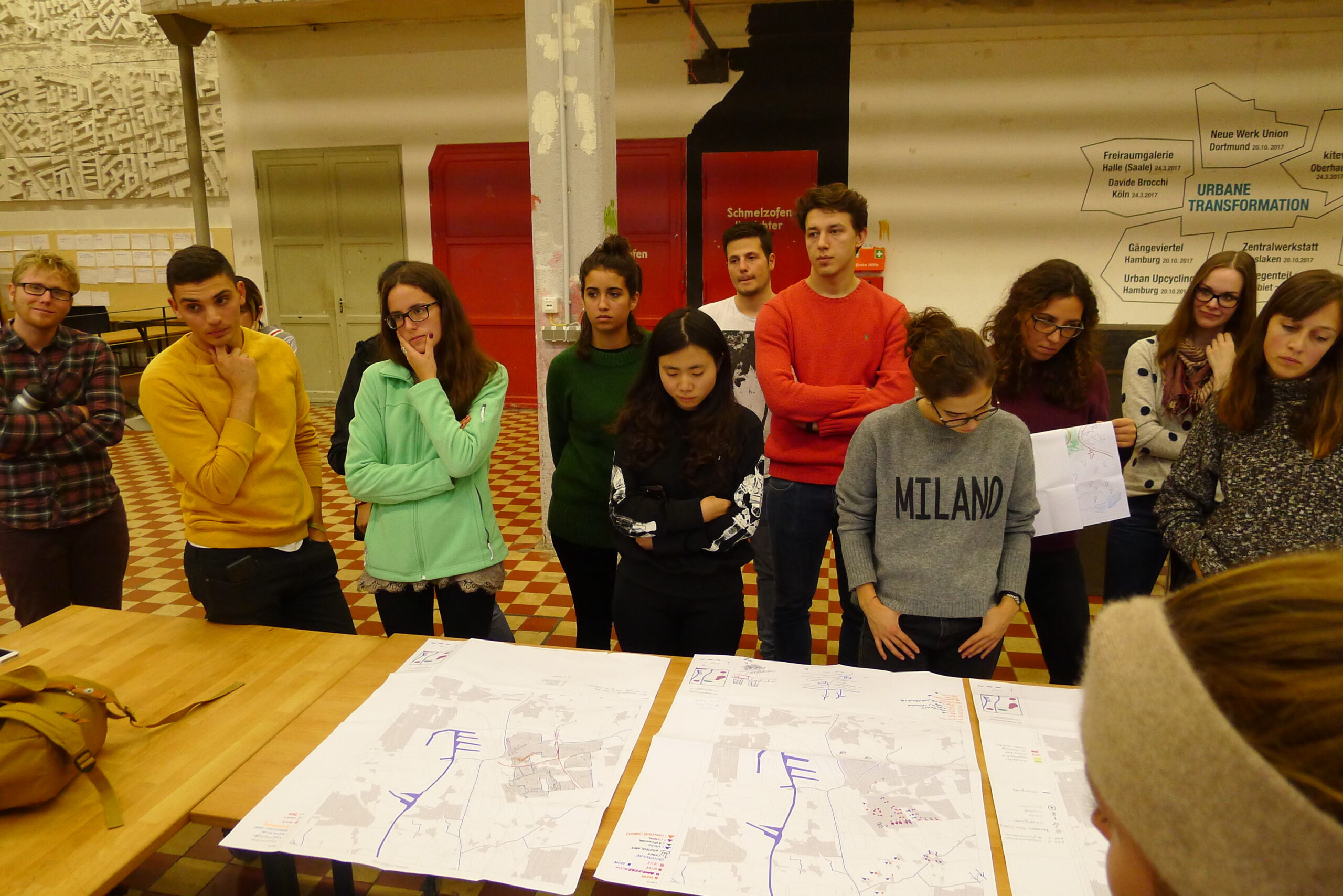 Eine Gruppe von etwa 15 Personen präsentiert Stadtplanungsskizzen auf Holztischen in einem Veranstaltungsraum.
