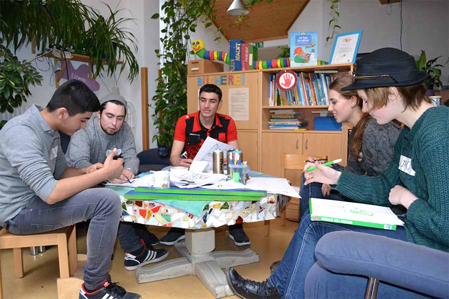 Sechs Personen sitzen in einem Kreis an einem Tisch und diskutieren über Dokumente in einem Klassenzimmer.