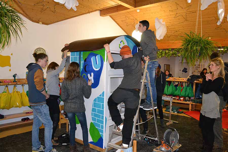 Mehrere Personen bemalen gemeinsam eine großformatige blaue Kunstinstallation in einer Halle.