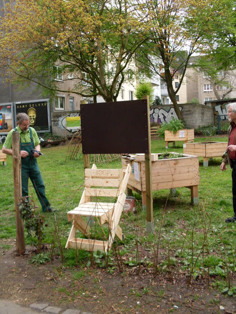 Gemeinschaftsgarten mit Hochbeeten, Treppen und Informationstafel, zwei Personen vor Wohngebäuden.
