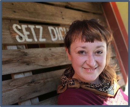 Eine Frau lehnt an einer Holzwand mit Aufschrift SETZ DICH, lächelt in die Kamera.