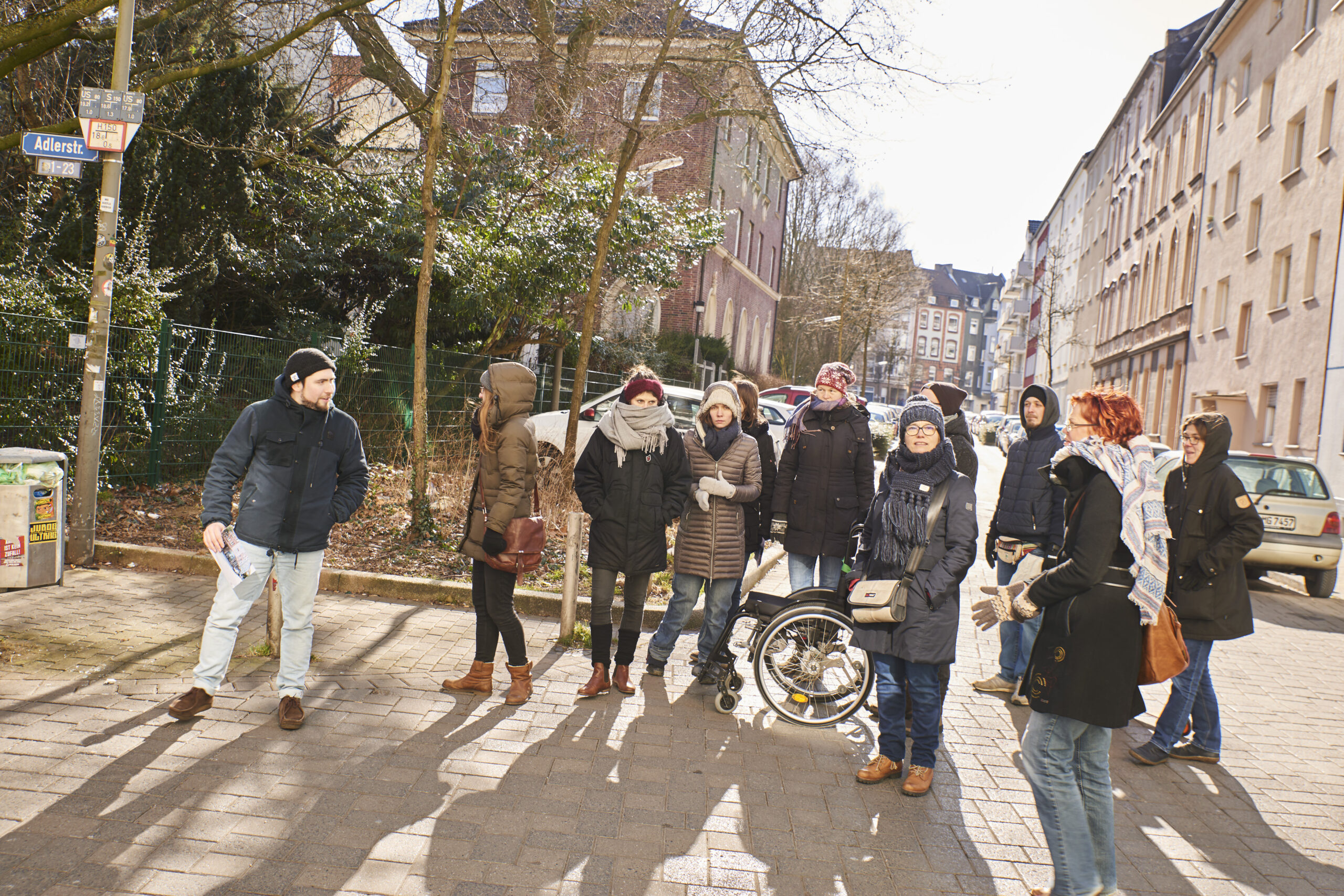 Eine Gruppe von etwa 15 Personen steht auf einem Gehweg in einer urbanen Wohnstraße zusammen.