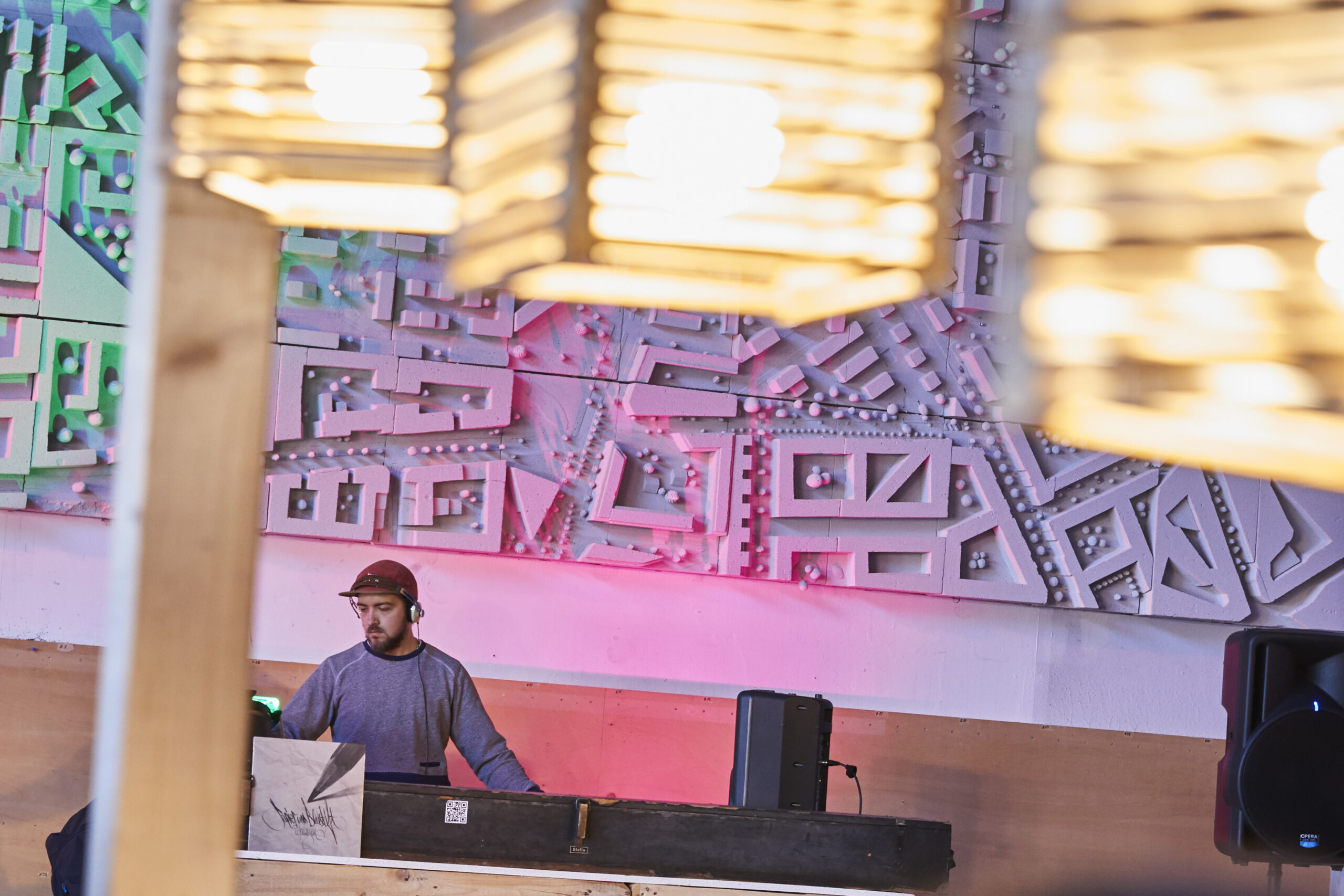 Mann an DJ-Pult vor großem Wandrelief mit architektonischen Mustern und Beleuchtung.