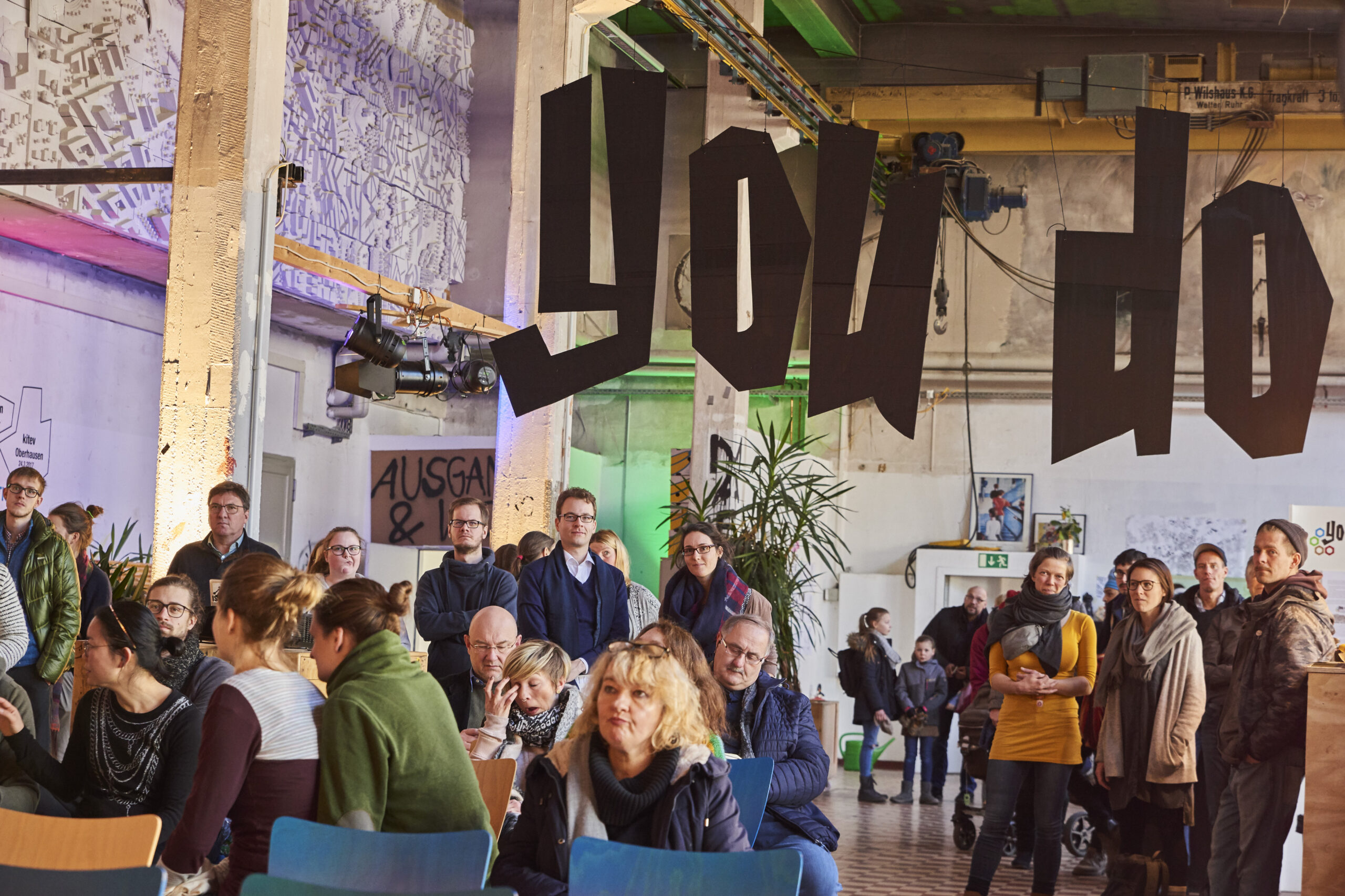 Etwa 50 Personen versammeln sich in einem industriellen Raum zu einer Veranstaltung oder Diskussion.