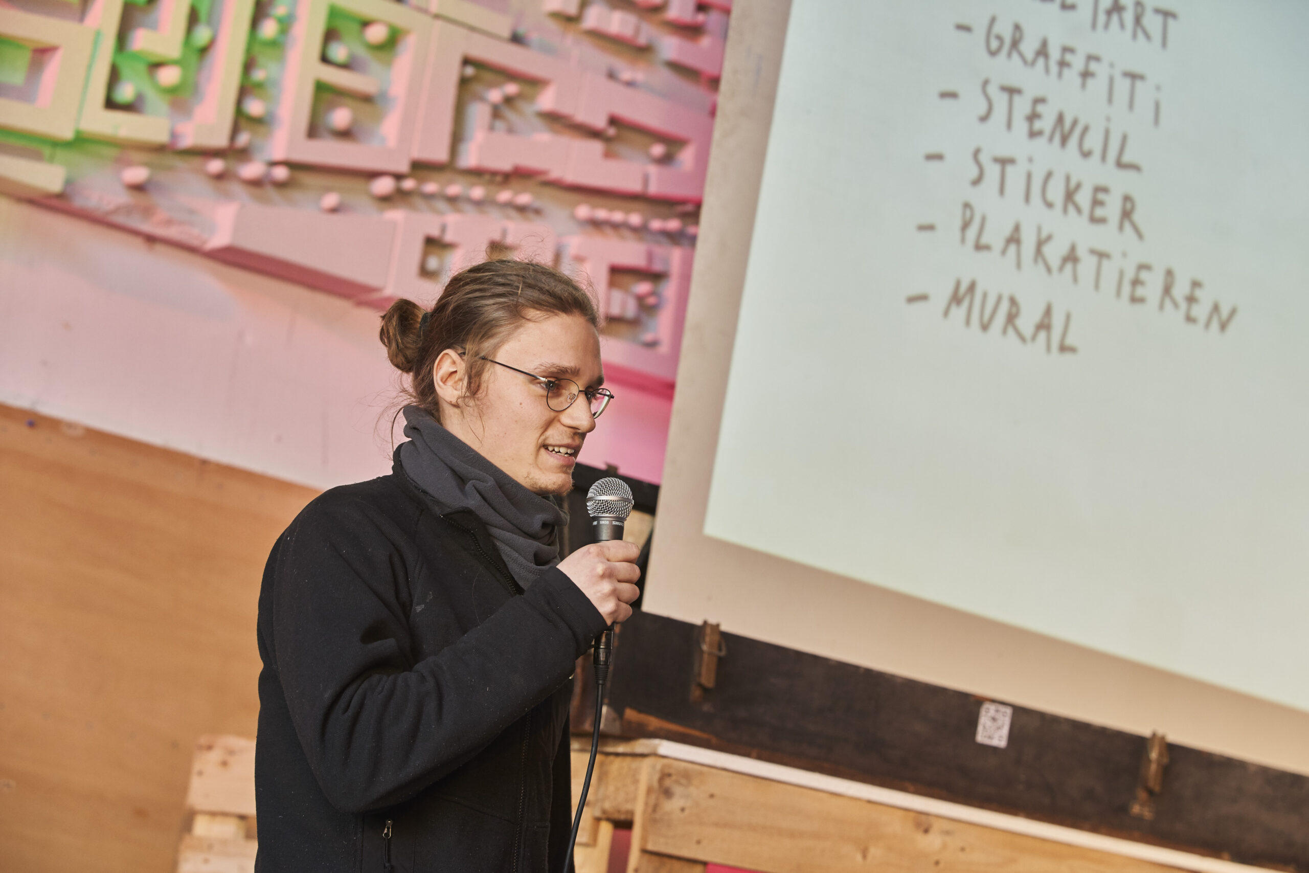 Eine Person mit Brille und schwarzer Jacke spricht mit Mikrofon vor einer Präsentationswand.