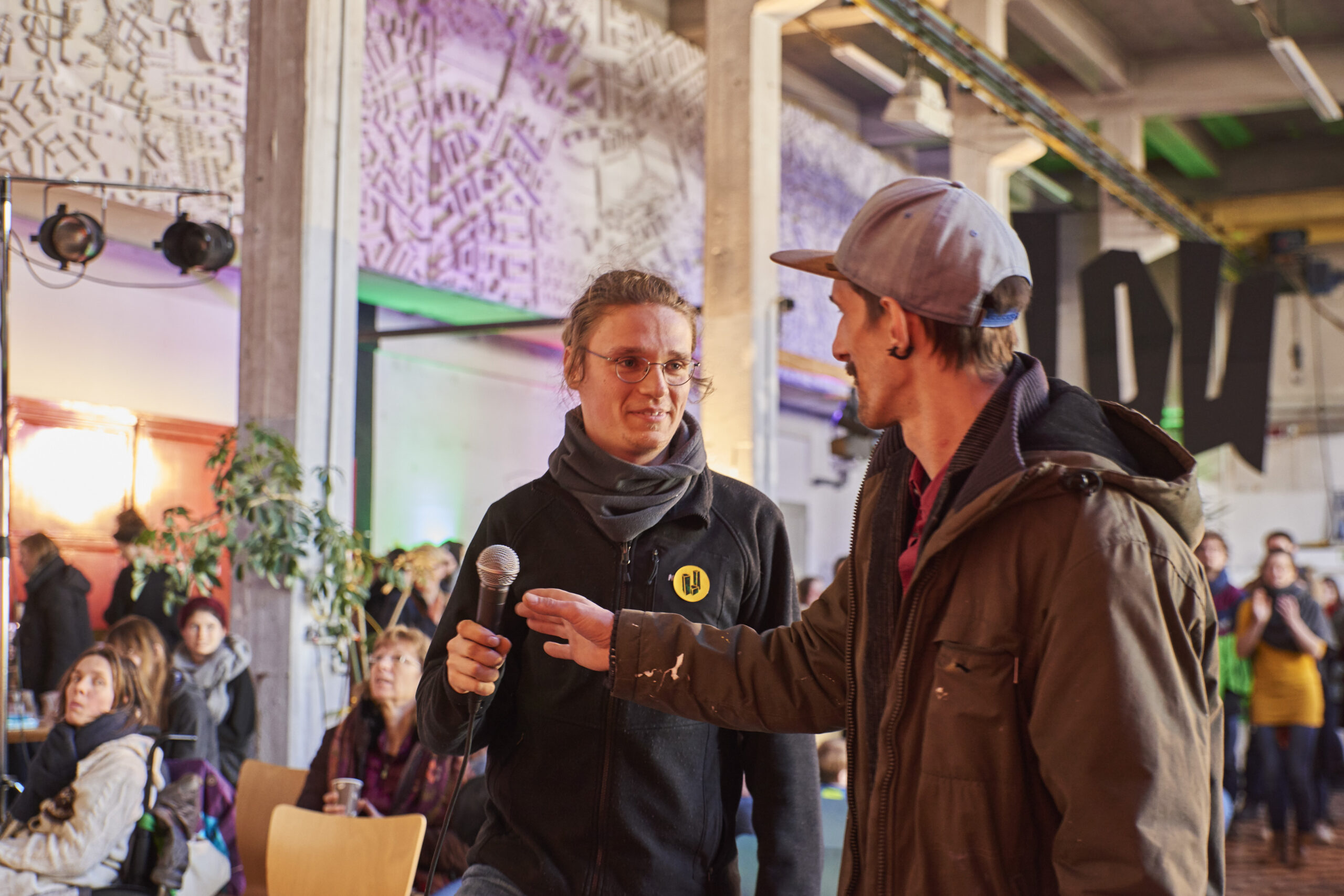 Zwei Männer diskutieren vor Publikum in einem Veranstaltungsraum mit Stadtplan-Projektion.