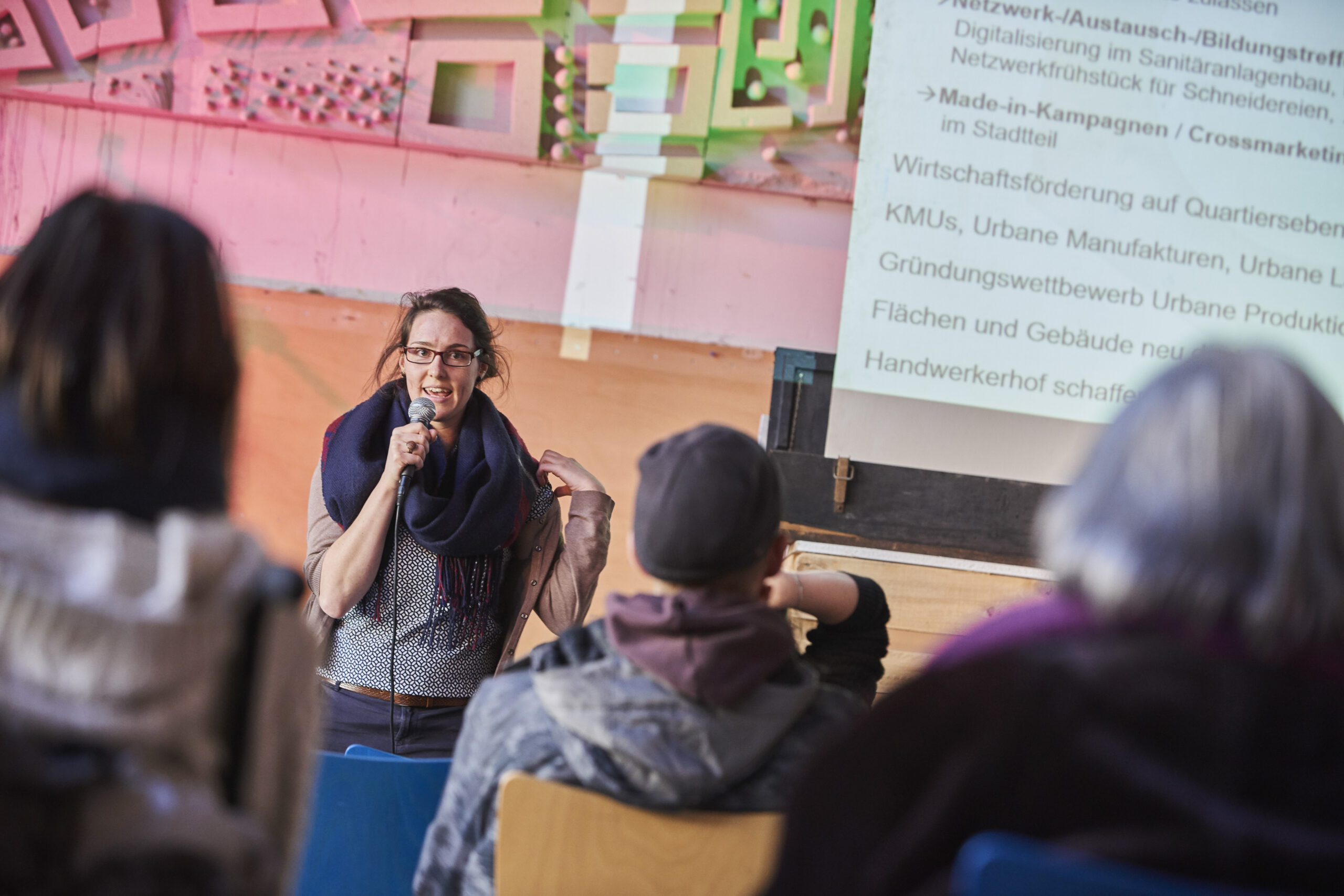 Eine Frau präsentiert mit Mikrofon vor Publikum ein Stadtentwicklungsprojekt an der Leinwand.
