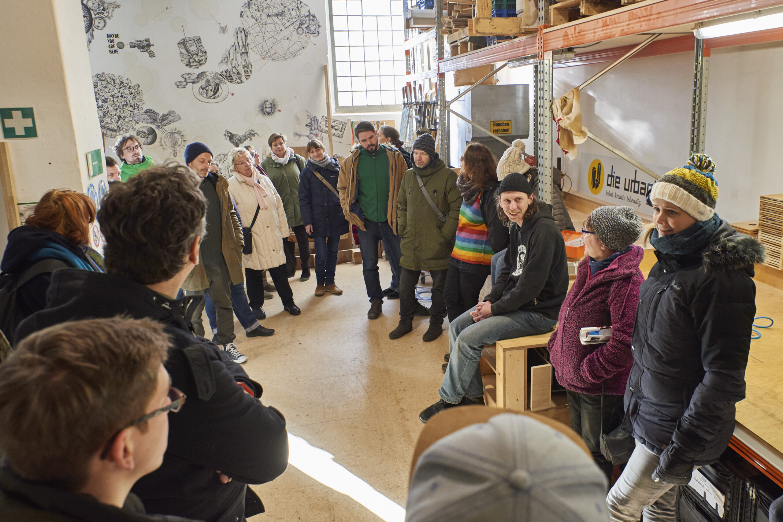 Eine Gruppe von etwa 25 Personen steht in einem Werkstatt-ähnlichen Raum im Kreis zusammen.