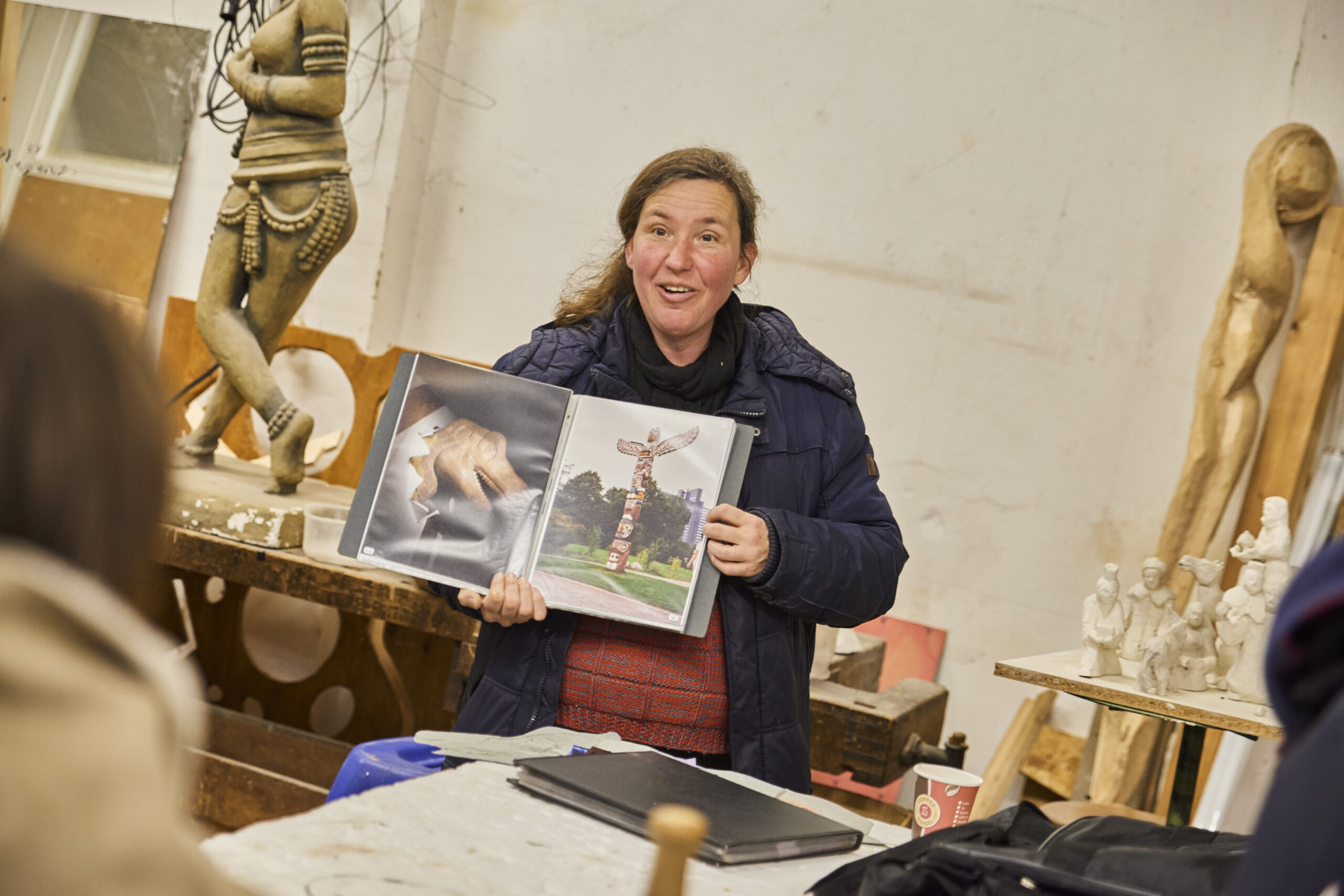 Frau präsentiert in Werkstatt mit Holzskulpturen Fotos von Kunstwerken und Totem-Pfosten.