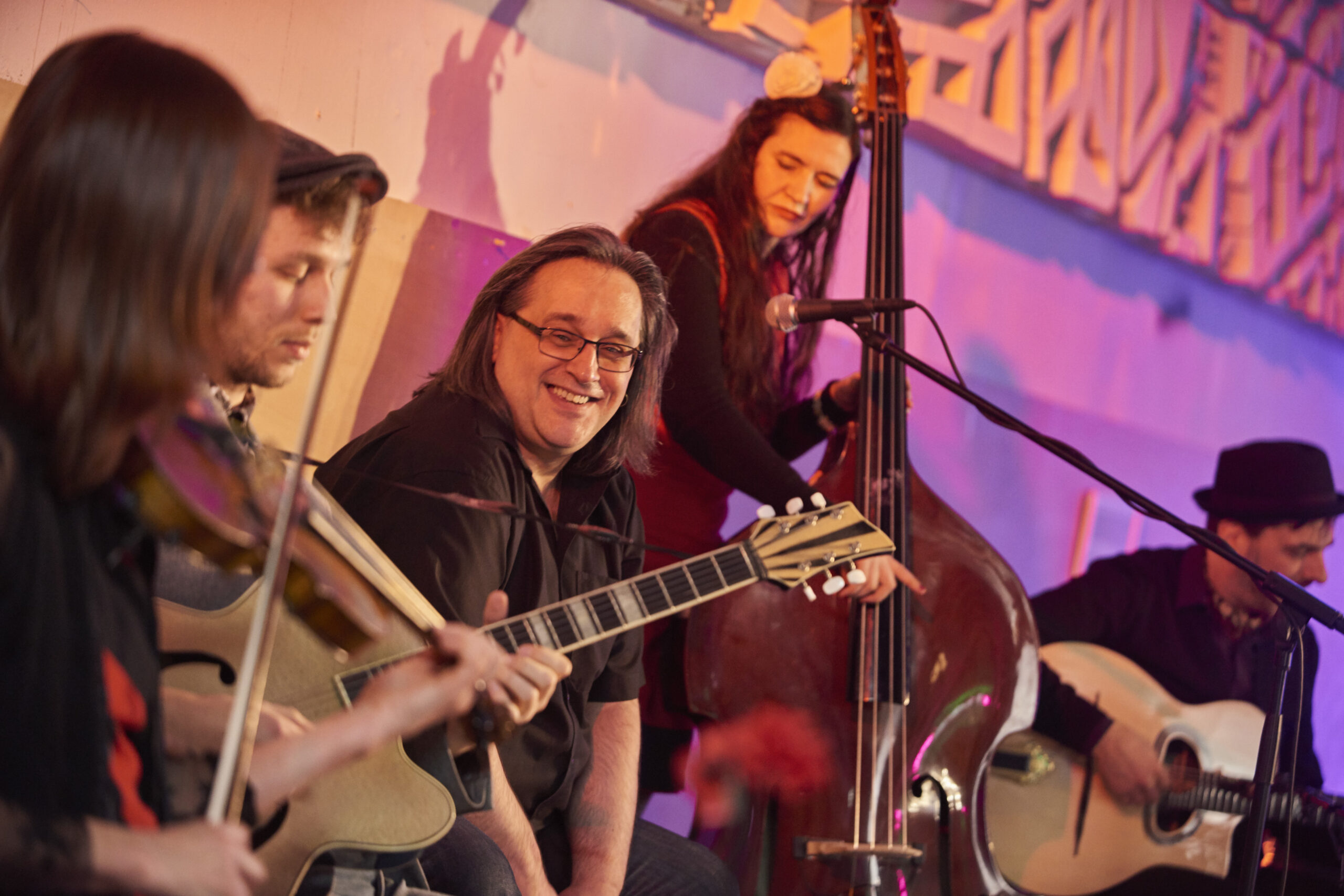 Vier Musiker spielen gemeinsam auf einer Bühne mit violetter Beleuchtung im Hintergrund.