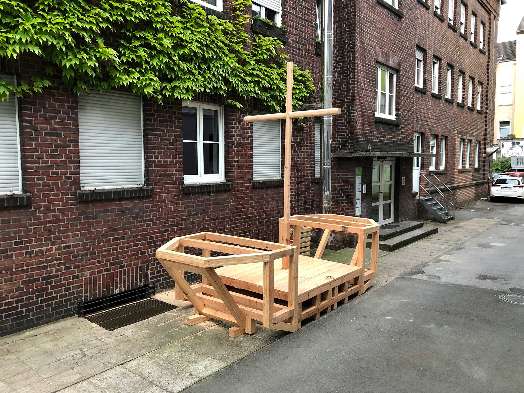Holzkonstruktion eines Bootes mit Kreuz in urbaner Hofeinfahrt zwischen Backsteinhäusern.