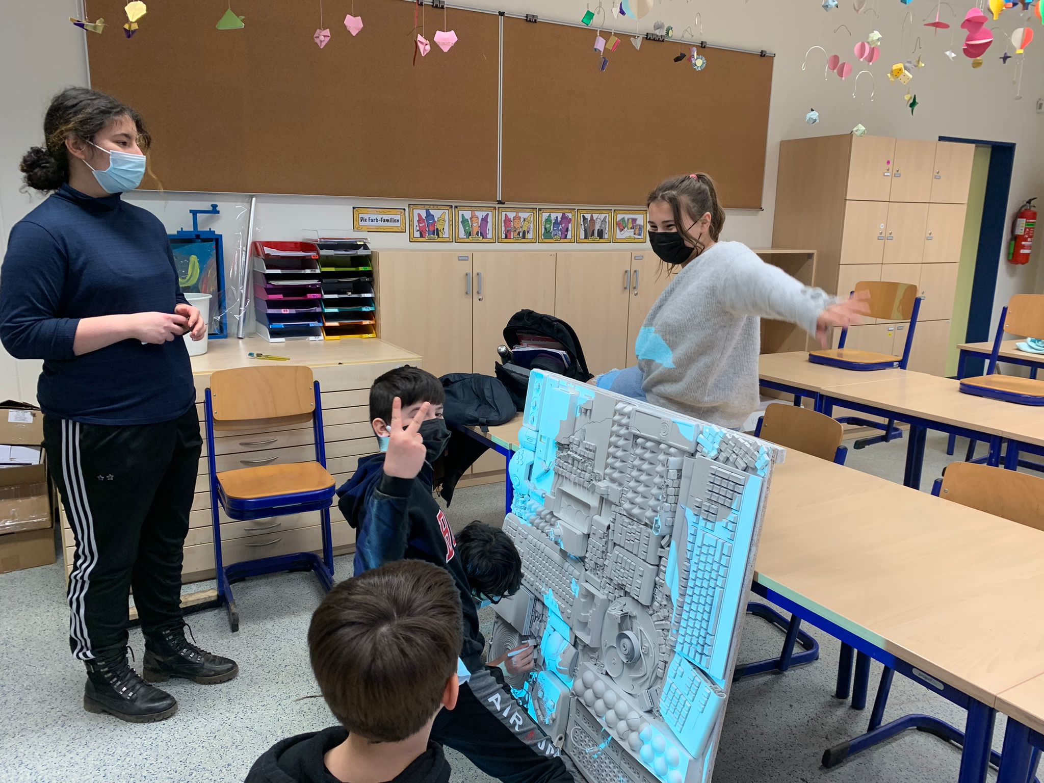 Zwei Erwachsene präsentieren Kindern ein großes blaues Architekturmodell in einem Klassenzimmer.