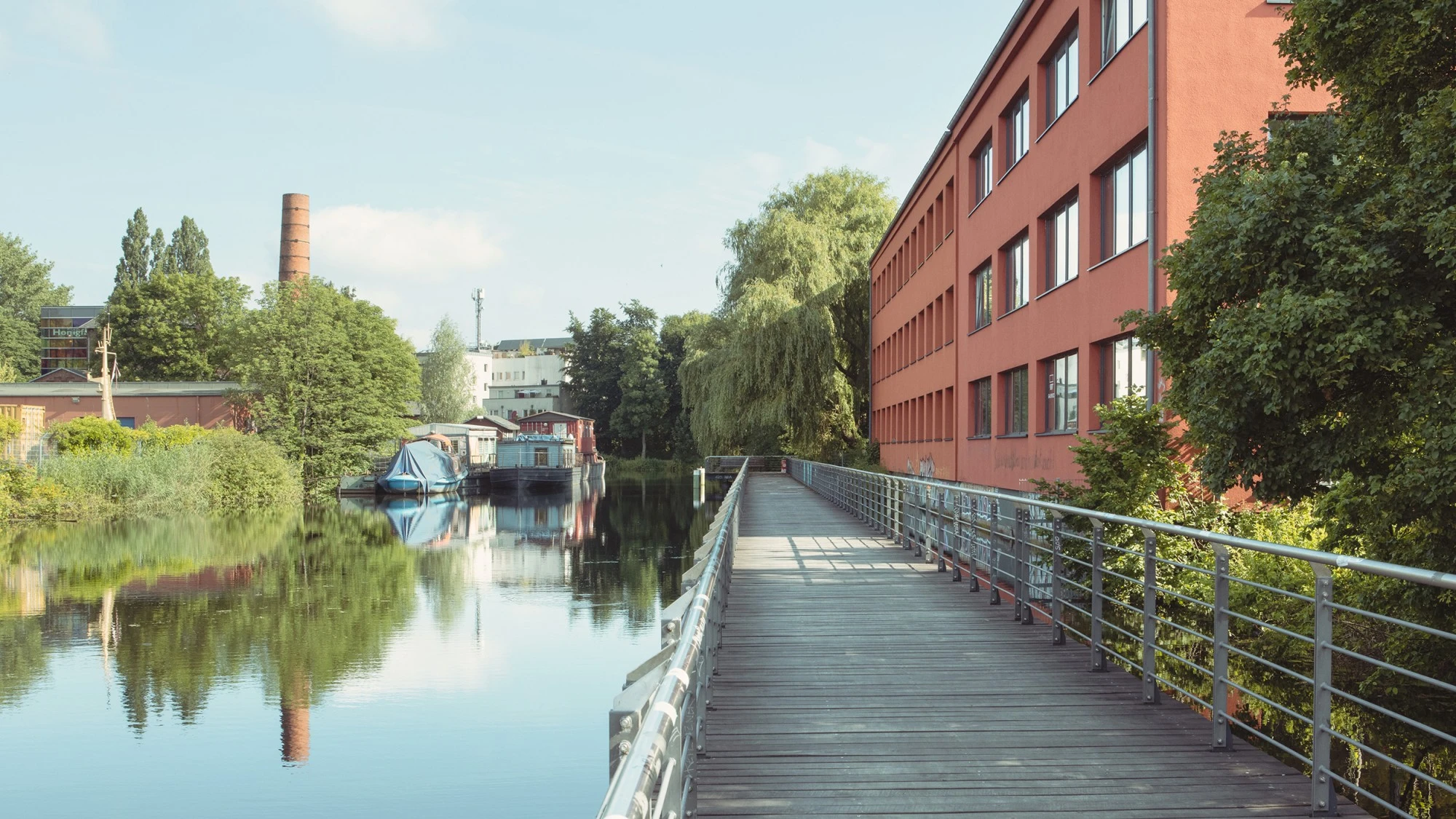 Holzsteg entlang eines Kanals mit Industriegebäuden, Booten und Ufervegetation.