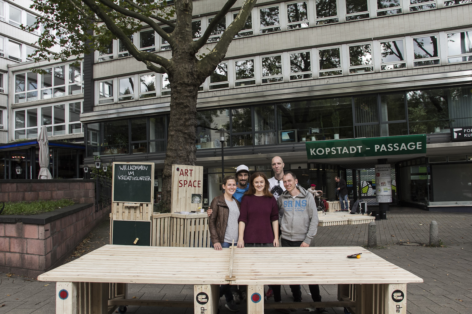 Fünf Personen stehen hinter einem großen Holztisch auf einer urbanen Platzfläche vor Geschäftshäusern.