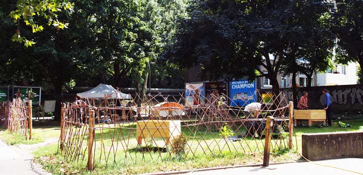 Improviser Marktplatz mit Holzstrukturen, Zelten und Menschen unter Bäumen in urbaner Umgebung.