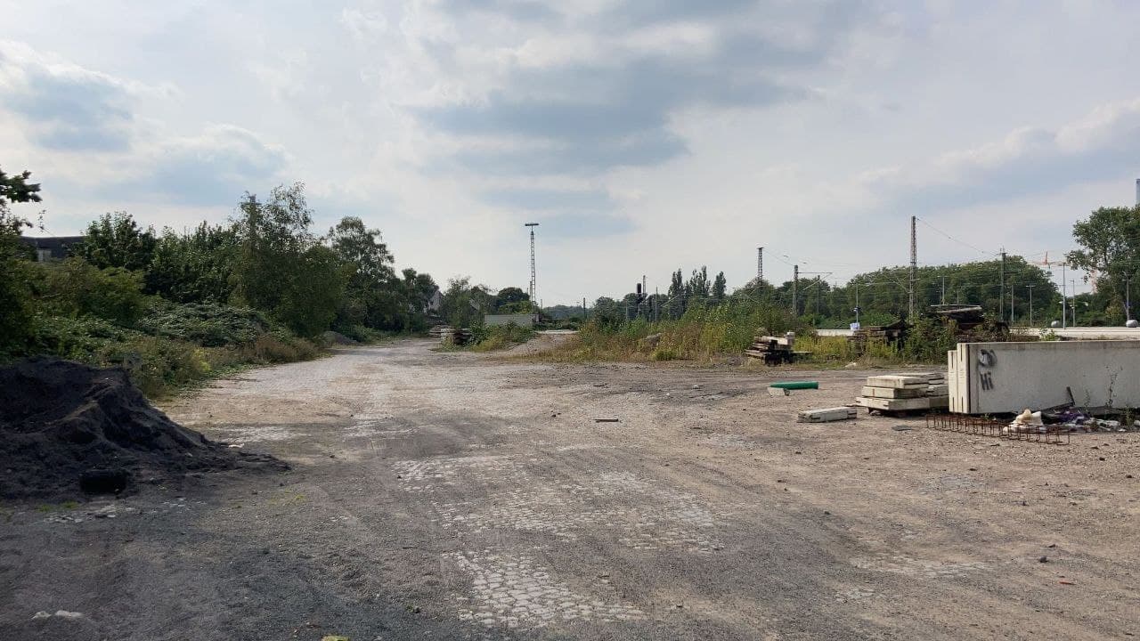 Brachliegende Industriefläche mit Gebäuden, Stromleitungen und umgebendem Grün.