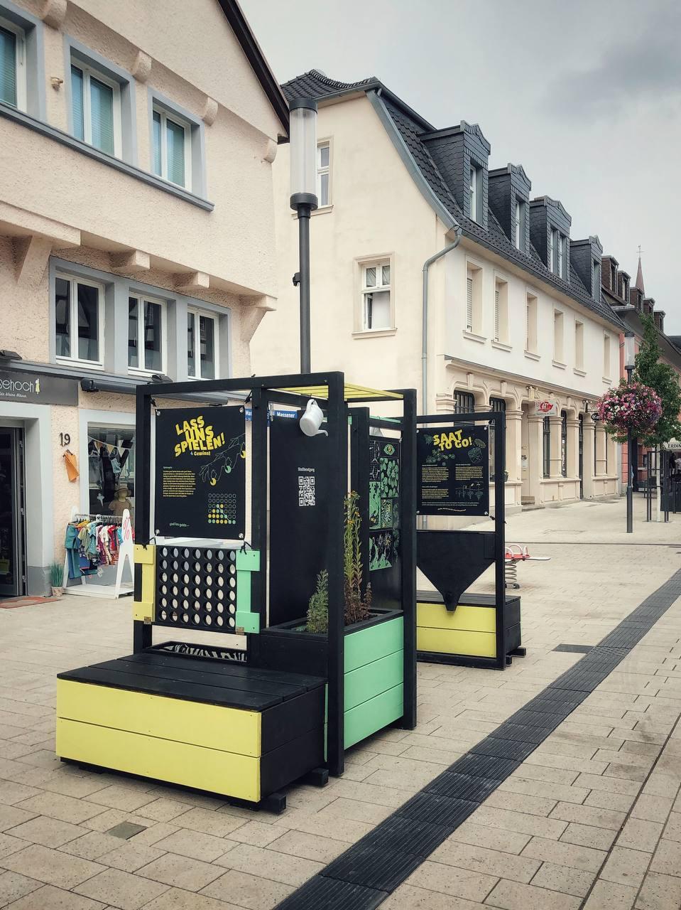 Interaktive Ausstellungsboxen mit schwarzem Rahmen und gelb-grünen Elementen auf Fußgängerzone.