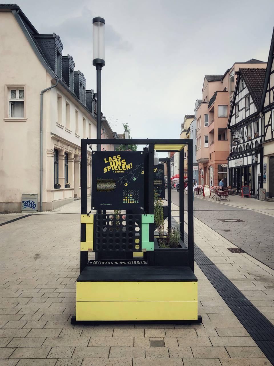 Installierte schwarze Kiosk-Struktur mit gelben und grünen Elementen in Fußgängerzone zwischen historischen Gebäuden.