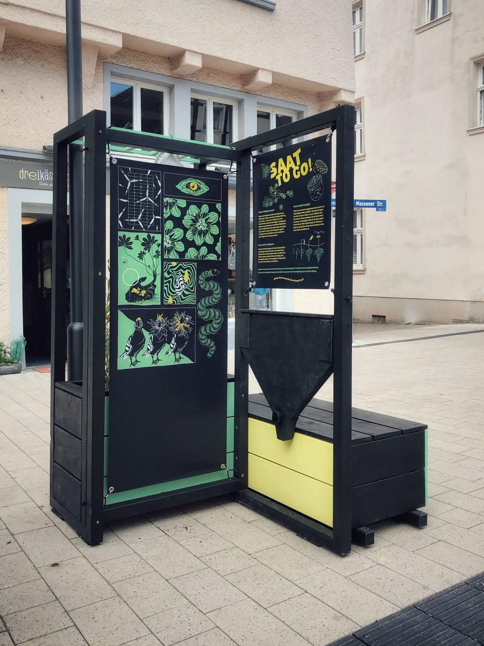 Schwarze Informationsbox mit grünen und gelben Elementen auf Gehweg vor Wohngebäude.