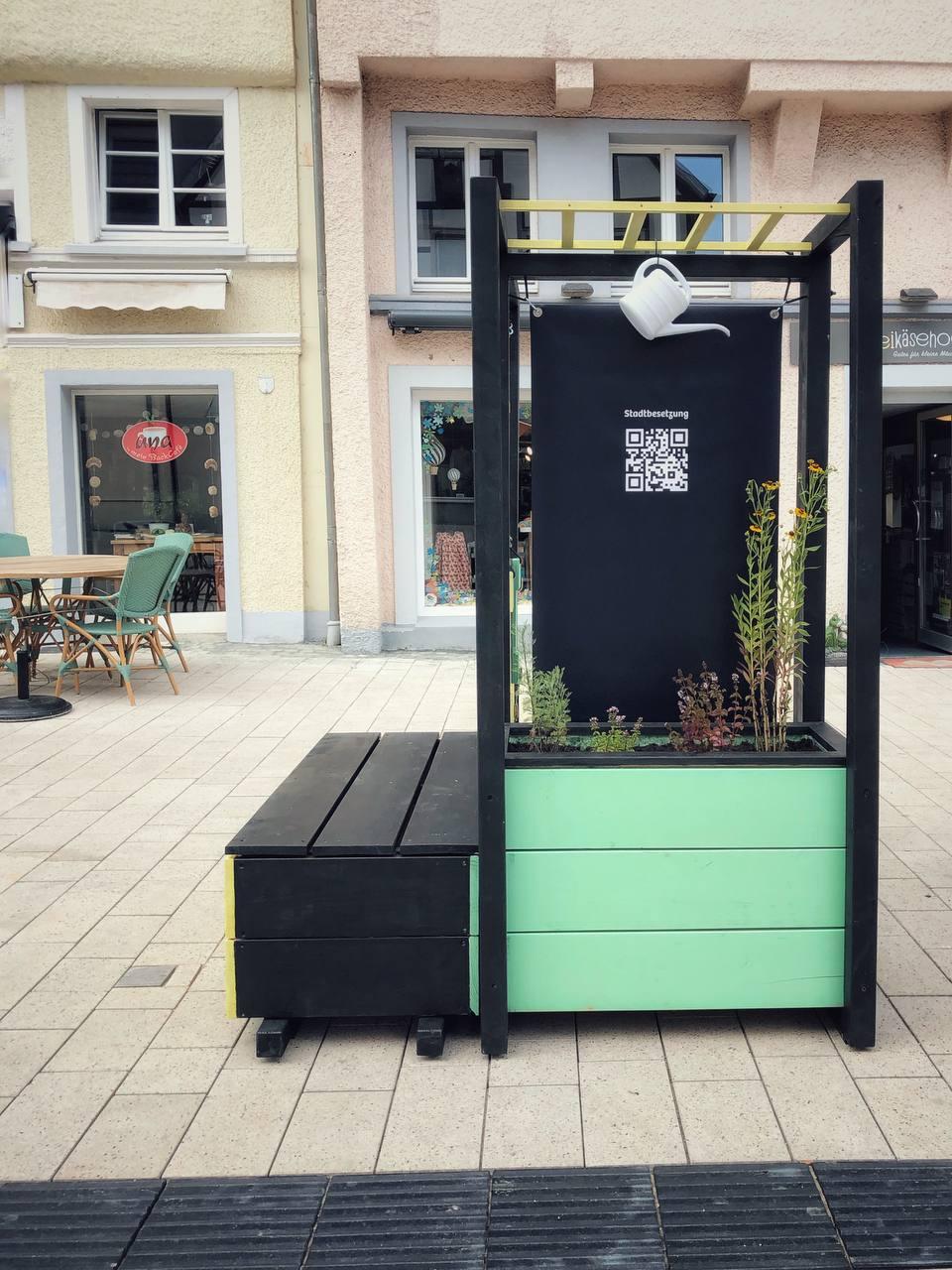 Moderne schwarze Stadtmöbel mit Begrünung und QR-Code auf belebtem Platz vor Gebäuden.