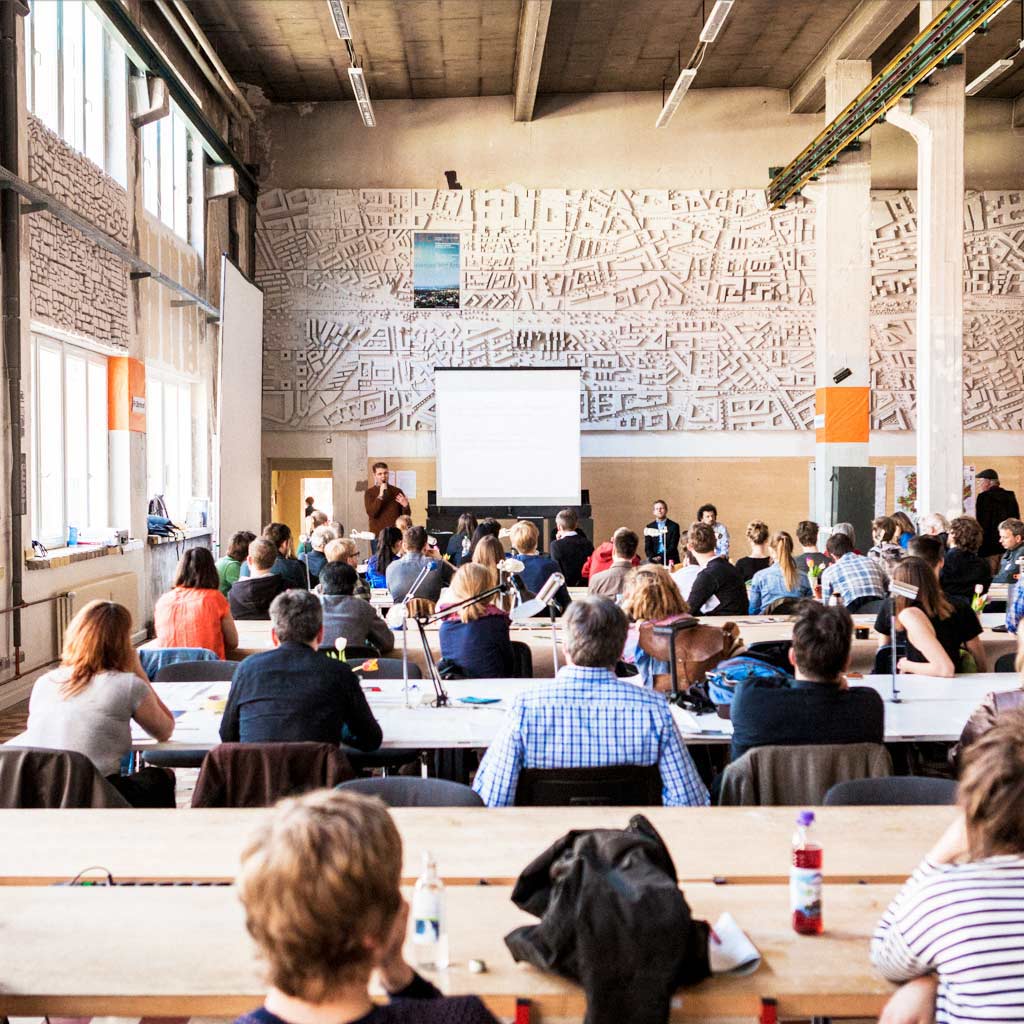 Großveranstaltung in Industriehalle mit Publikum an Tischen und Projektionsfläche an Wand.