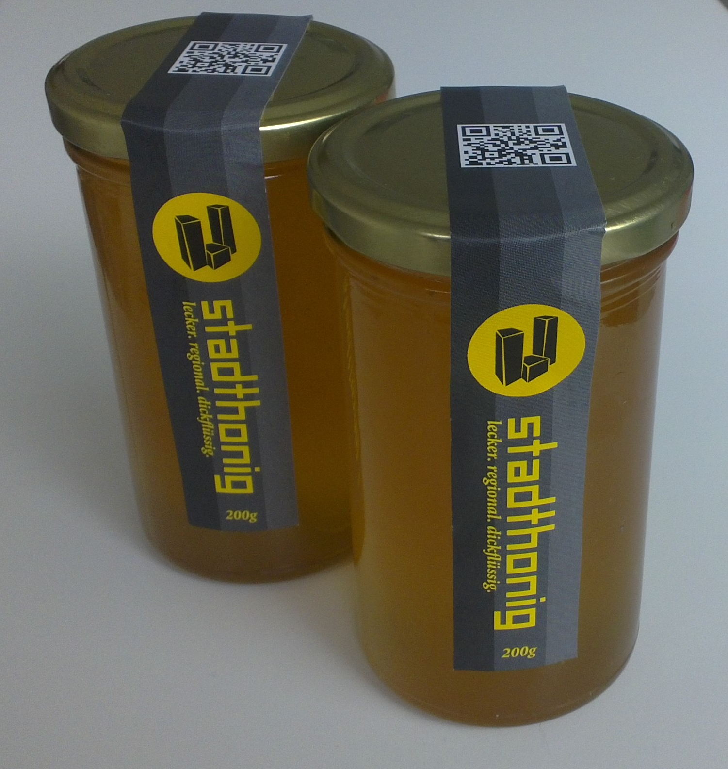 Zwei Gläser mit honigfarbenem Inhalt, schwarze Bänder mit gelbem Logo und QR-Code.