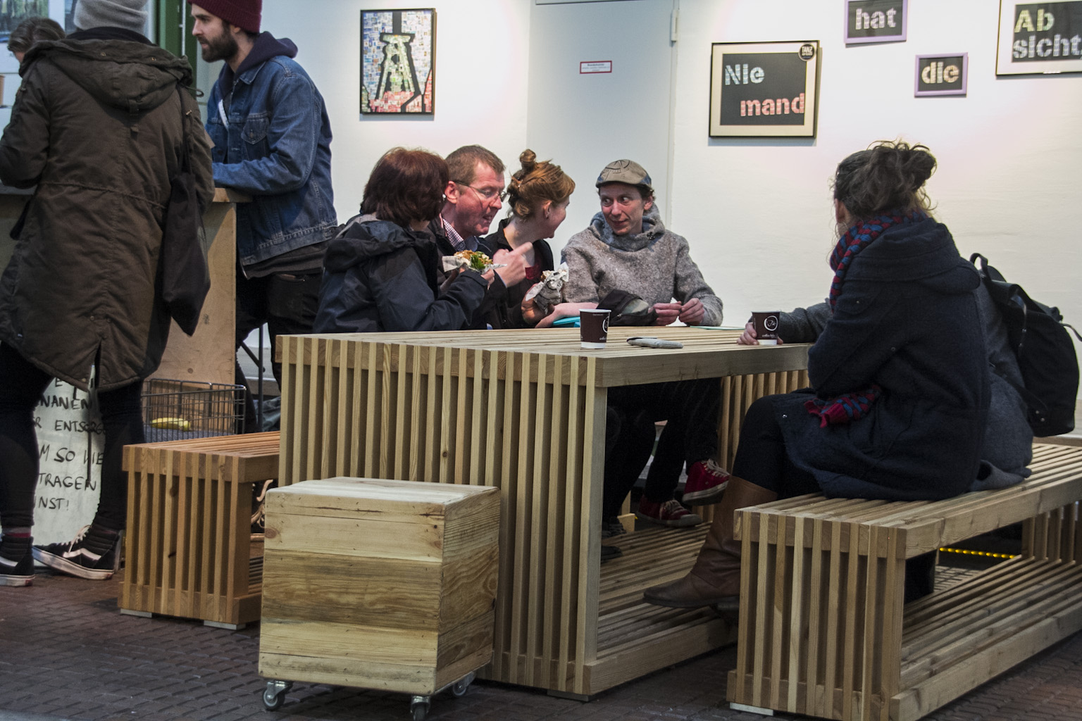 Mehrere Personen sitzen an einem Holztisch in einem Raum mit deutschen Textplakaten an den Wänden.