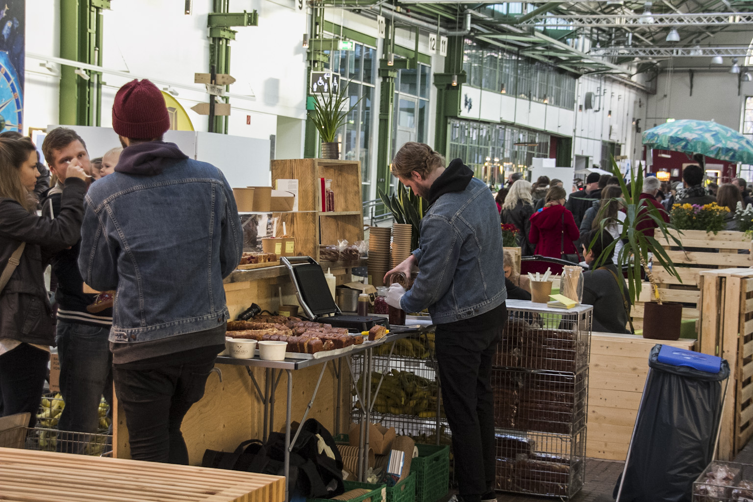 Besucher kaufen Speisen an einem Stand auf einem Markt in einer Industriehalle.