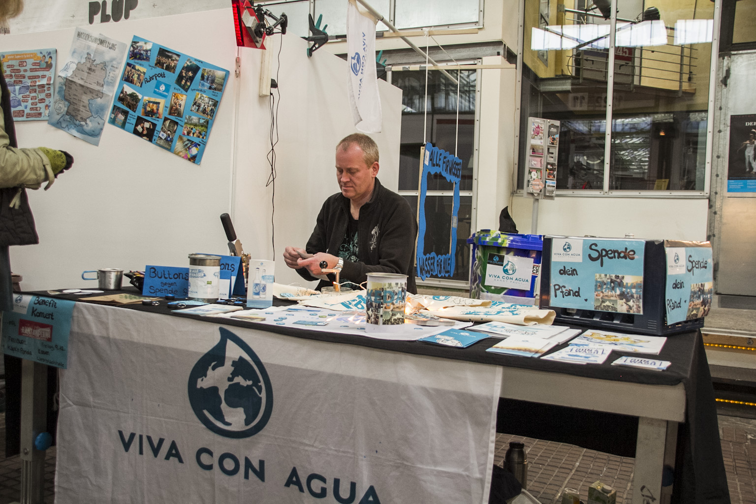 Mann sitzt an Informationsstand einer Organisation namens Viva Con Agua hinter Tisch.