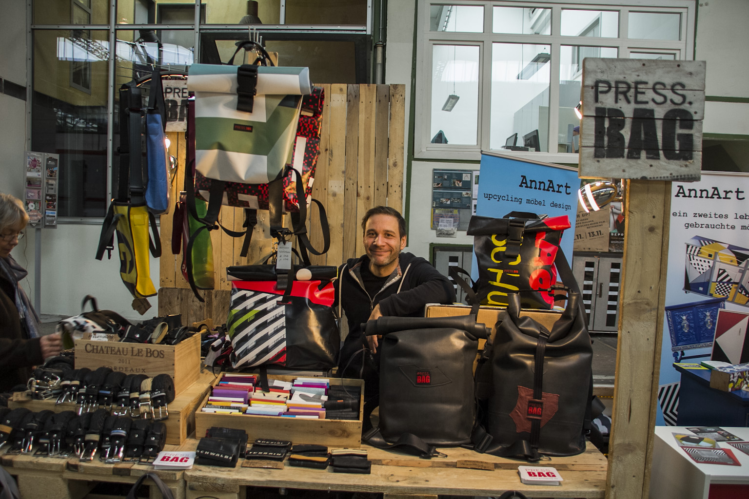 Mann sitzt an Marktstand mit selbstgestalteten Taschen und Rucksäcken aus recycelten Materialien.