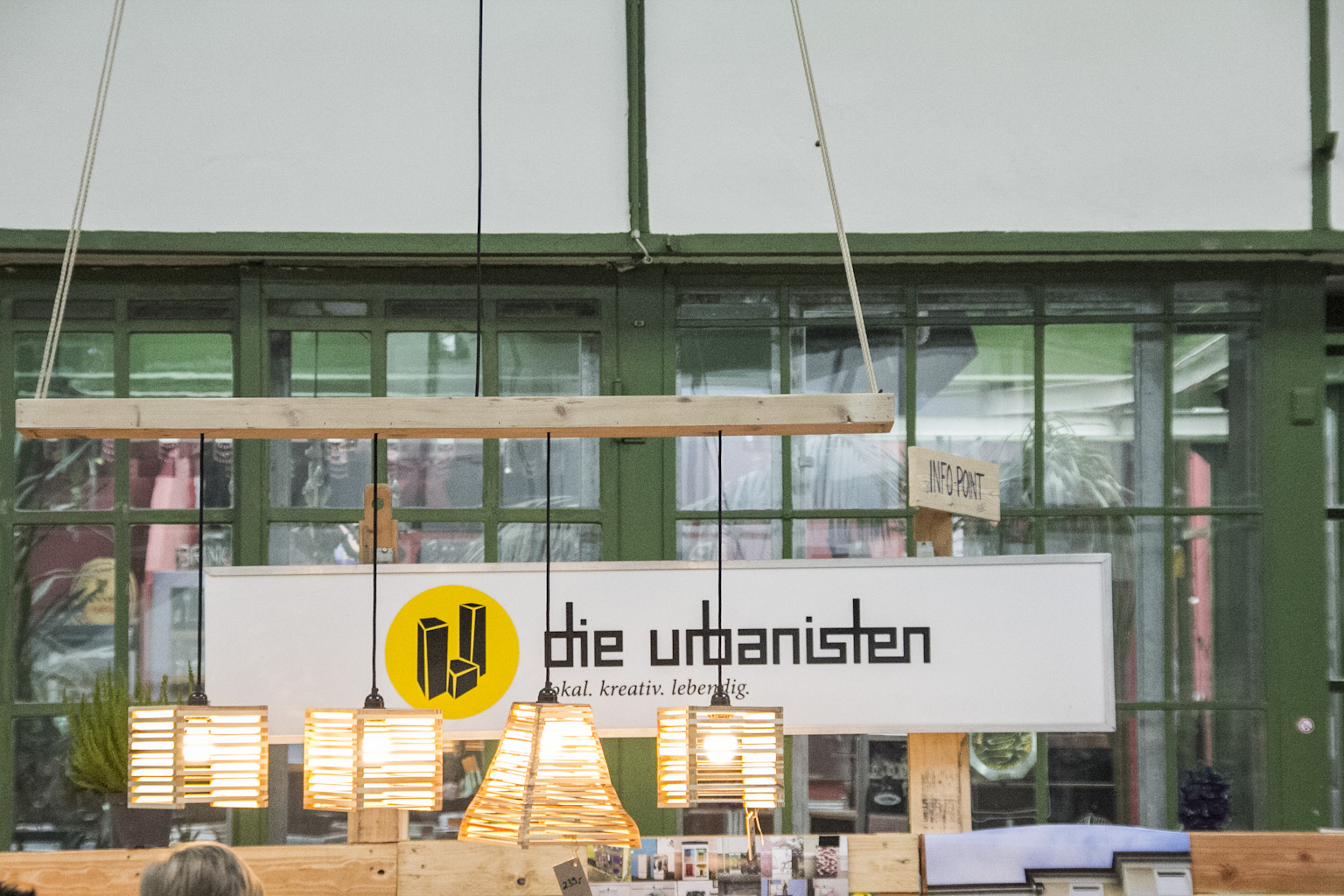 Storefront mit grünen Rahmen, Logo und Schriftzug die urbanisten, hängenden Holzleuchten.