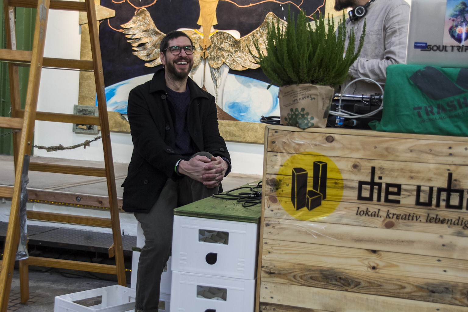 Mann mit Brille sitzt lächelnd in Werkstatt neben Holzschild mit die Urbis-Logo.