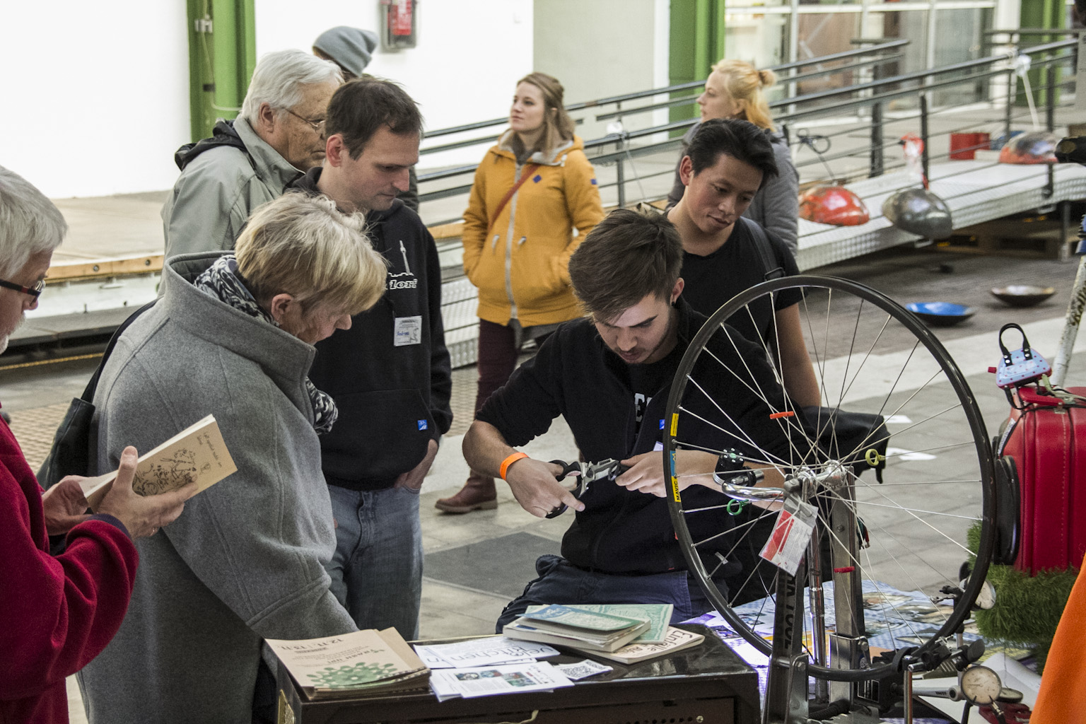Eine Person erklärt einer Gruppe Besuchern ein Fahrradrad bei einer Veranstaltung in einer Halle.
