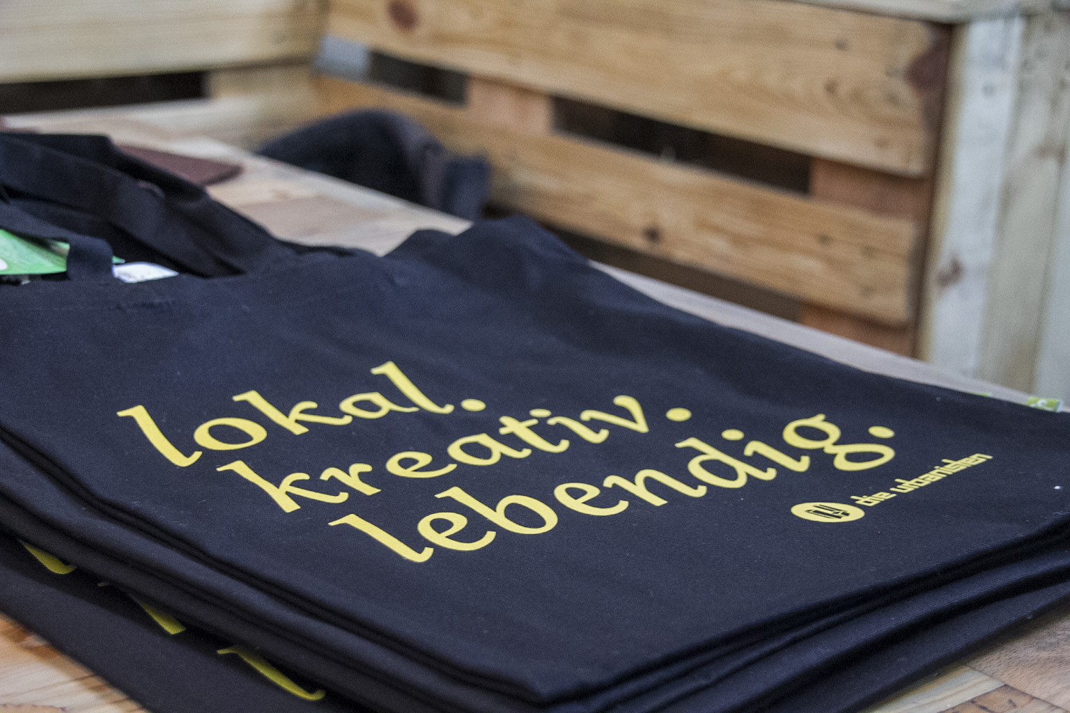 Stapel dunkler T-Shirts mit gelbem Aufdruck lokal kreativ lebending auf Holztisch.