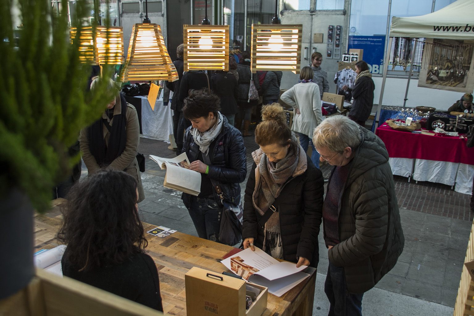 Besucher betrachten Ausstellungsmaterialien an Tischen in einer Halle mit Designleuchten.