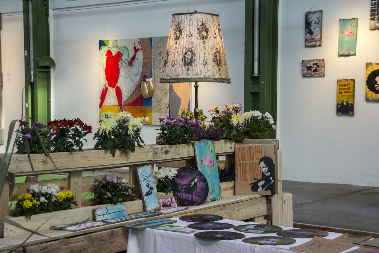 Ausstellungsraum mit Holzgestellen, Blumenschmuck, Lampe und an Wänden aufgehängten Kunstwerken.