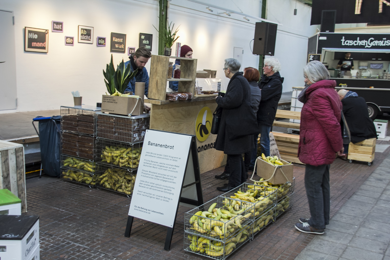 Ein Marktstand mit Bananenbrot wird von Besuchern besucht, mehrere Menschen stehen wartend in einer Schlange.