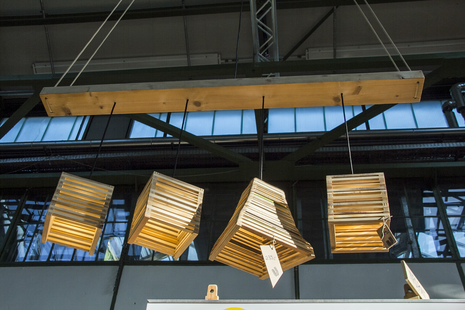 Vier hölzerne Lampen mit Lamellenschirmen hängen an einem Balken in einer Industriehalle.
