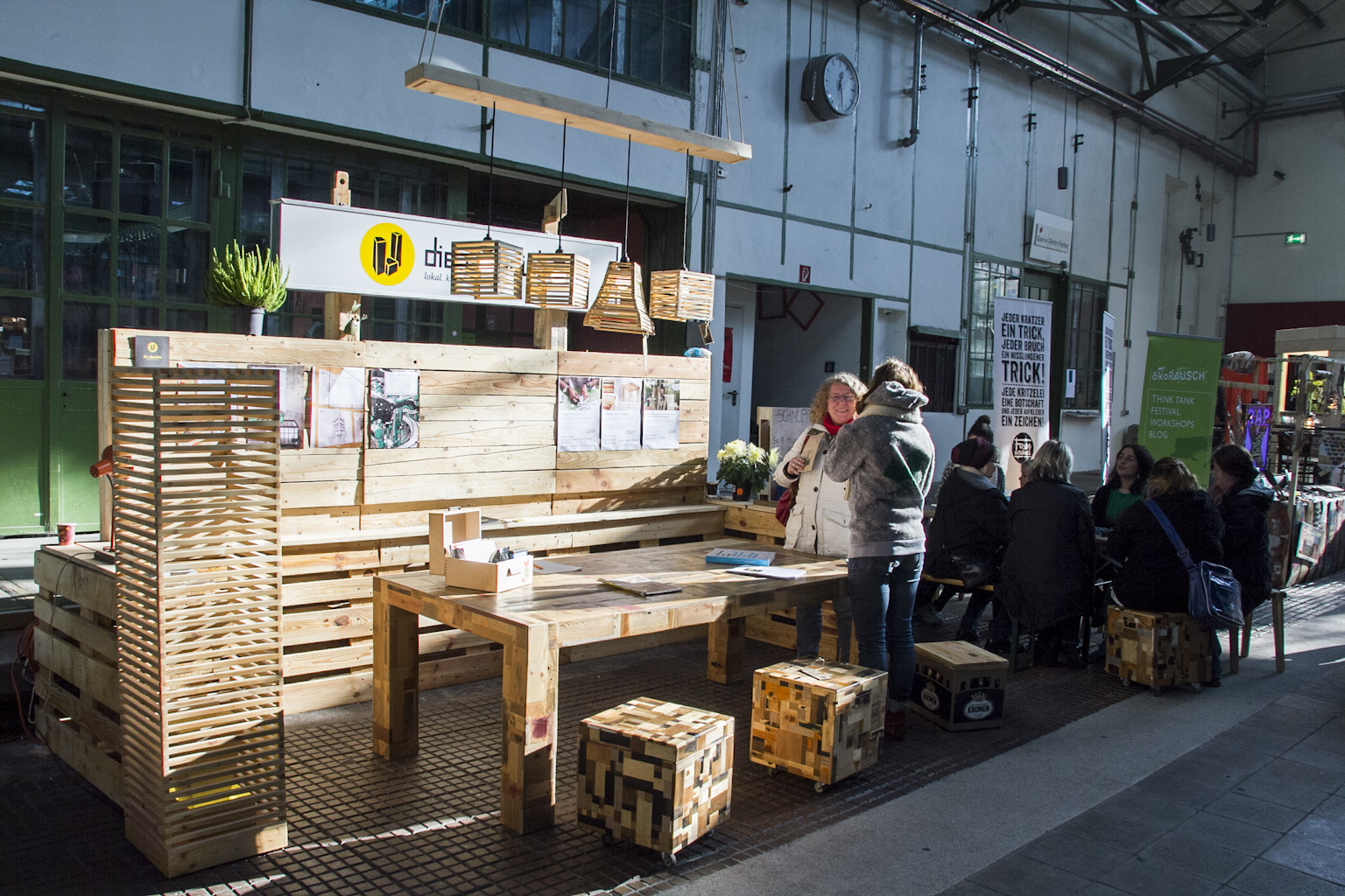 Holzkonstruktion mit Sitzbereichen und Hängeleuchten in einer Indoor-Markthalle mit Besuchern.